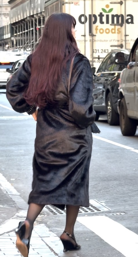 Black faux fur coat with long burgundy hair and black wedge heels walking on a city street.
