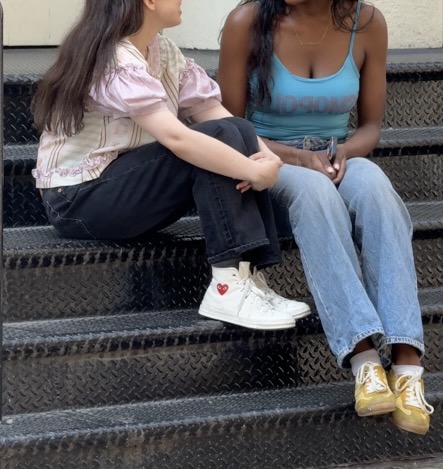 Two women on stairs. One wears a striped top and black jeans, the other a blue top and jeans.