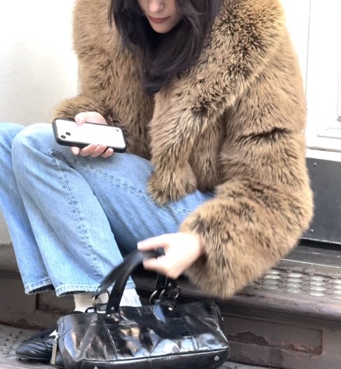 Brown faux fur jacket with light wash jeans and black shiny handbag.