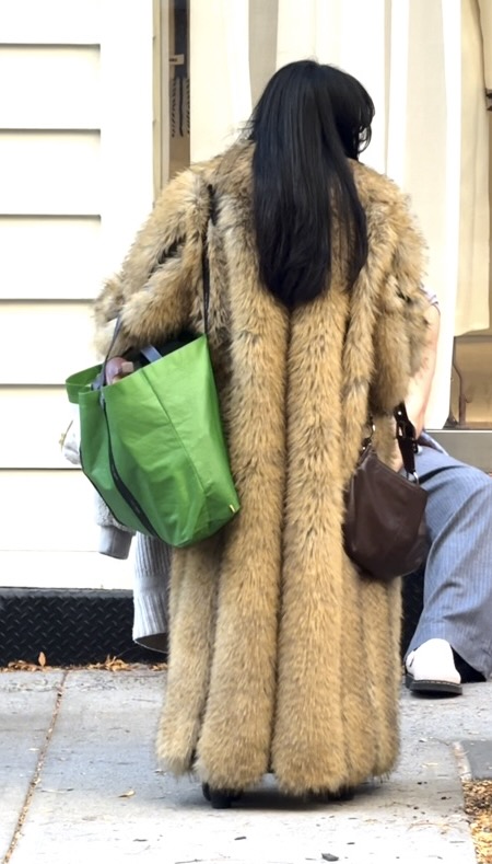 Long tan fur coat with black hair, carrying a green tote and brown purse.
