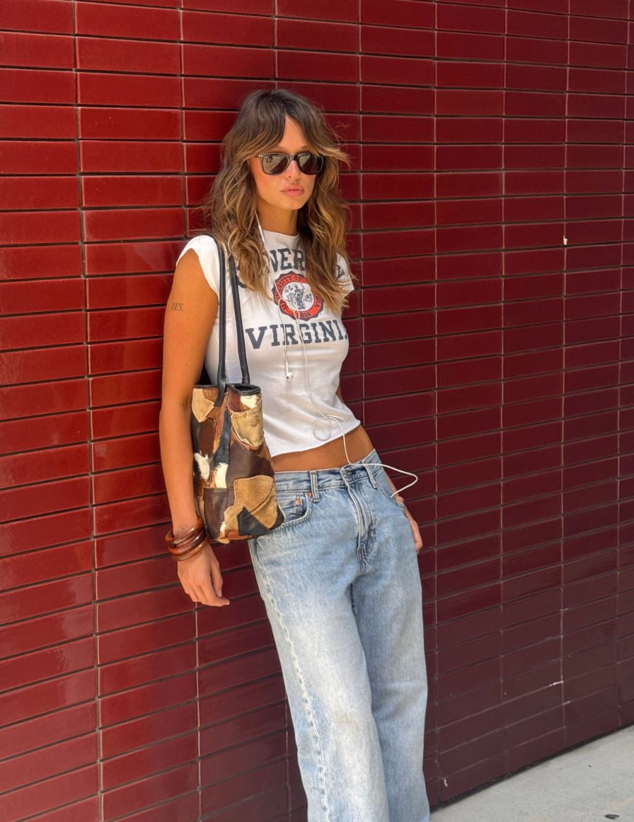 White graphic tee, light wash jeans, patchwork bag, and sunglasses against a red wall.
