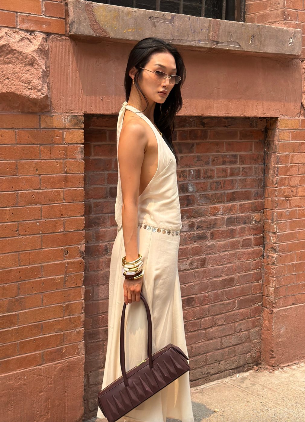 Cream halter maxi dress with coin belt, gold bangles, sunglasses, and a brown baguette bag.