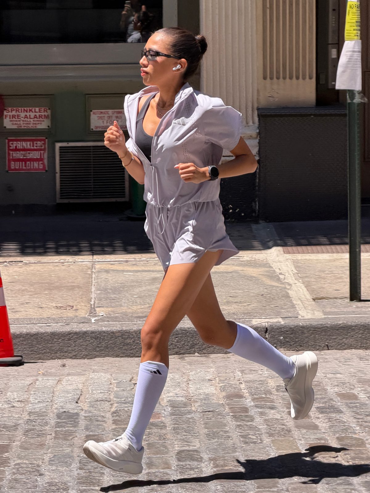 Light lavender hooded running romper with black sports bra, white socks, sneakers, and sunglasses.